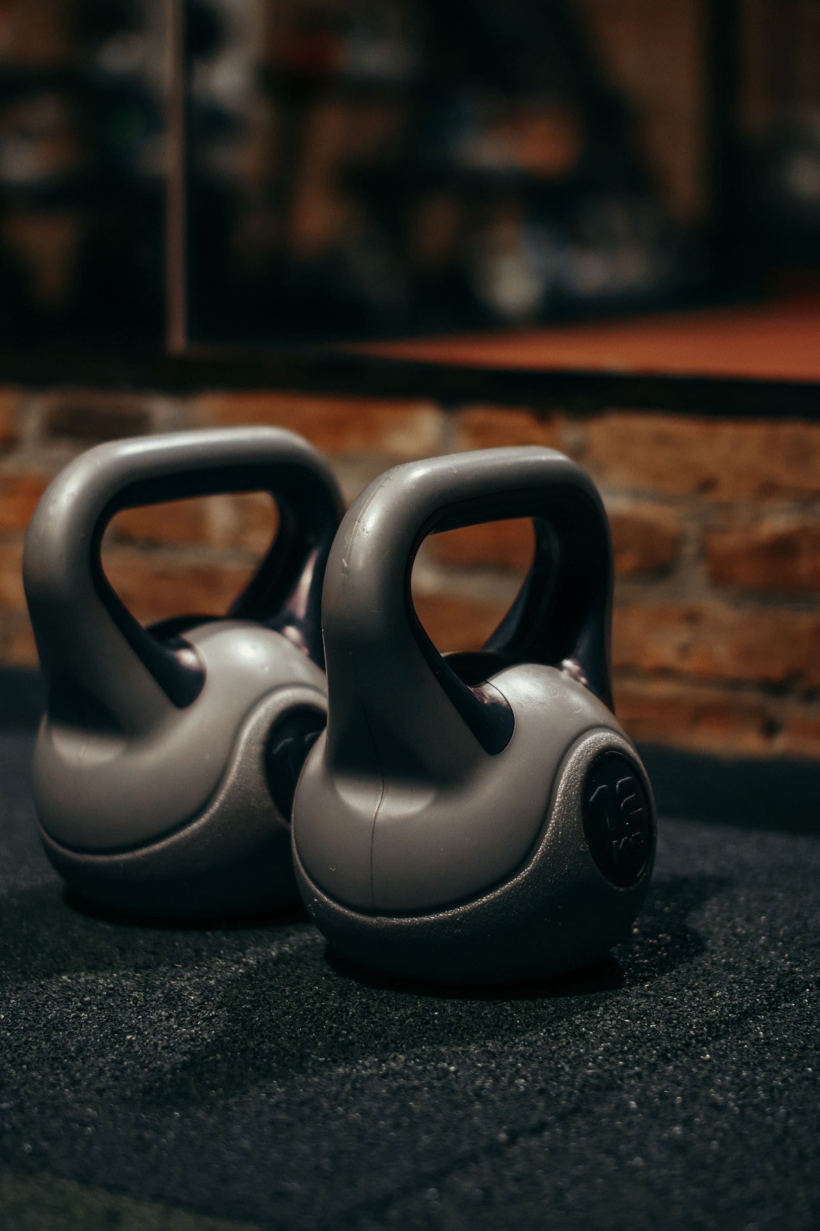 Two grey kettlebells on a rubber floor in front of a brick wall